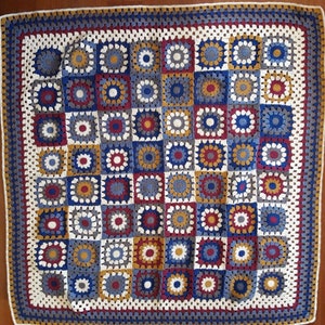 May include: A crocheted blanket with a square pattern in shades of blue, white, red, and gold. The blanket is made of yarn and has a fringed border.