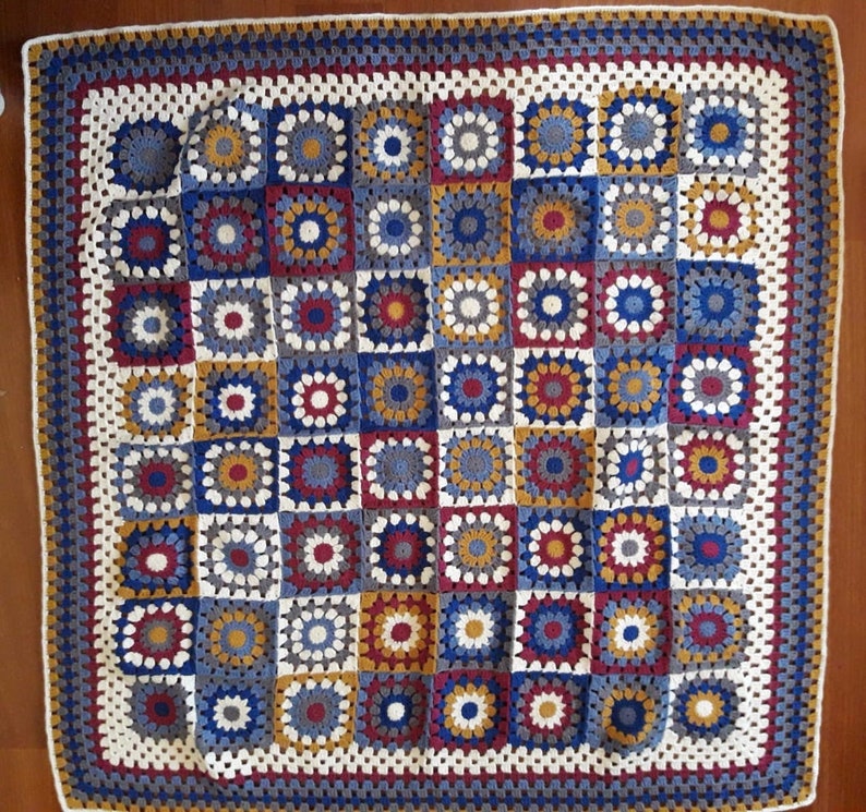 May include: A crocheted blanket with a square pattern in shades of blue, white, red, and gold. The blanket is made of yarn and has a fringed border.