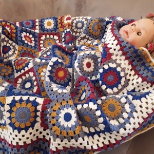 May include: A crocheted baby blanket with a colorful geometric pattern in shades of blue, red, white, and yellow. The blanket is made of soft yarn and has a delicate texture.