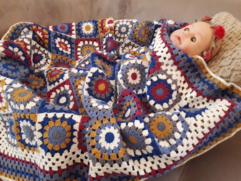 May include: A crocheted baby blanket with a colorful geometric pattern in shades of blue, red, white, and yellow. The blanket is made of soft yarn and has a delicate texture.