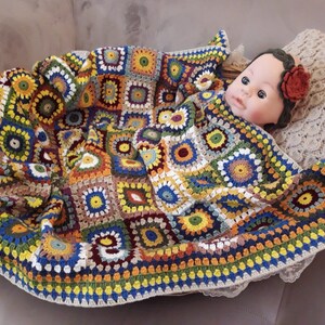 May include: A colorful crocheted baby blanket with a multi-colored granny square pattern. The blanket is made with a variety of yarn colors, including yellow, orange, red, green, blue, and brown. The blanket is folded and draped over a wicker basket, with a baby doll lying on top of the blanket.