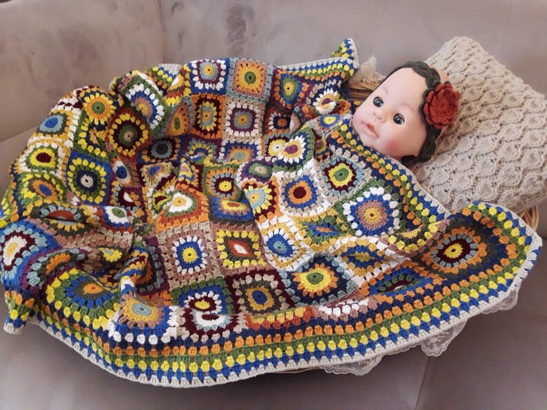 May include: A colorful crocheted baby blanket with a multi-colored granny square pattern. The blanket is made with a variety of yarn colors, including yellow, orange, red, green, blue, and brown. The blanket is folded and draped over a wicker basket, with a baby doll lying on top of the blanket.