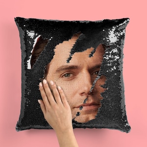 May include: A black sequin pillow with a man's face peeking out from a hole in the center. The face is visible from the forehead to the chin. The pillow is being held by a hand.