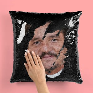 May include: A black sequin pillow with a man's face peeking out from the sequins. The man has dark hair, a beard, and is smiling.