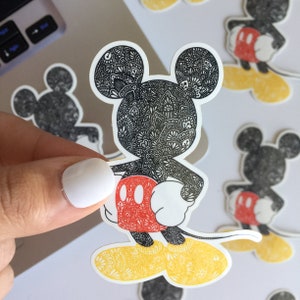 May include: A black and white patterned Mickey Mouse sticker with red shorts and yellow shoes. The sticker is on a white background.