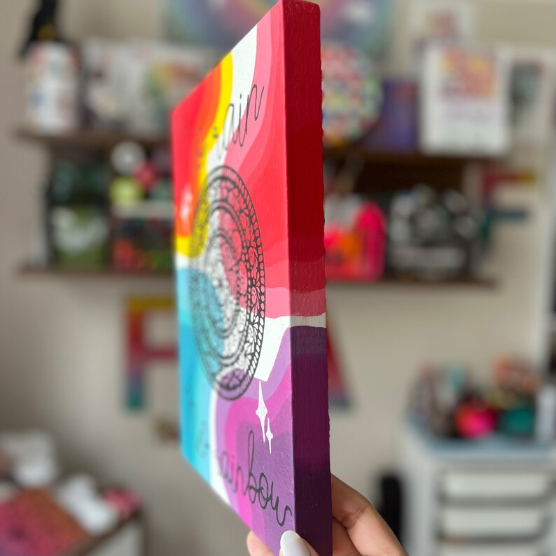 May include: A colorful canvas painting with a rainbow design and the text "The rain" and "rainbow" on the sides. The painting is on a wooden frame.