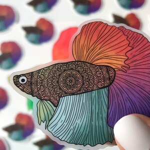 May include: A colorful sticker of a betta fish with a mandala pattern on its head. The fish has a red, orange, yellow, blue, and purple tail.