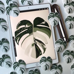 May include: A white tablet computer with a green and white monstera leaf design on the screen. The tablet is surrounded by several smaller monstera leaf stickers in various shades of green and white.