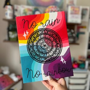 May include: A colorful canvas painting with a rainbow background and a black and white mandala in the center. The text "No rain no rainbow" is written in black cursive.