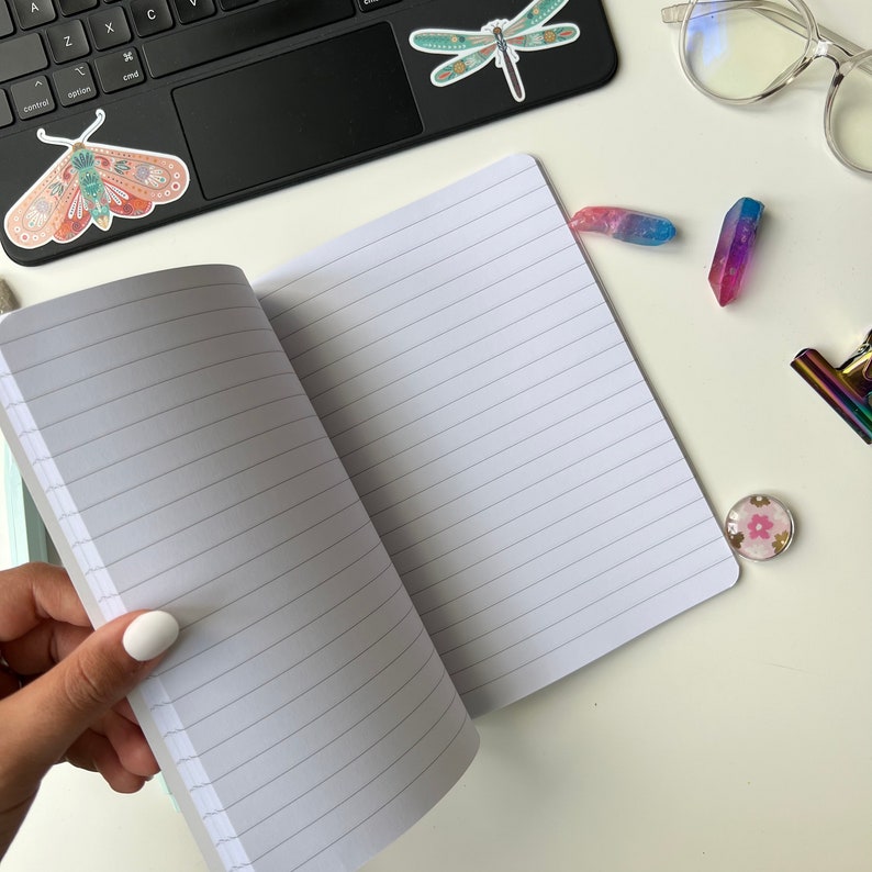 May include: An open lined notebook with a white background and blue lines. The notebook is on a white surface with a dragonfly sticker, a butterfly sticker, and a few colourful crystals.