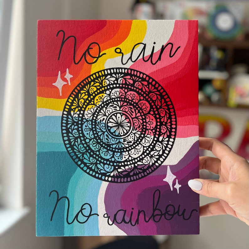 May include: A colorful painting with a rainbow background and a black and white mandala in the center. The text "no rain no rainbow" is written in black at the bottom of the painting.