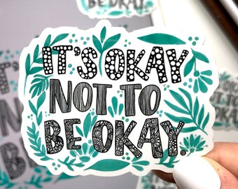 May include: A teal green sticker with black and white lettering that reads "It's okay not to be okay." The sticker has a floral design with leaves and flowers.