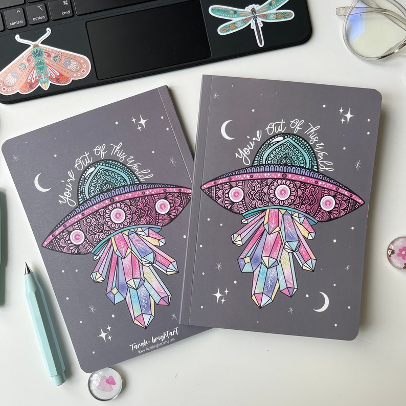 May include: Two grey notebooks featuring a colourful illustration of a UFO with crystals and the text "You're Out Of This World".
