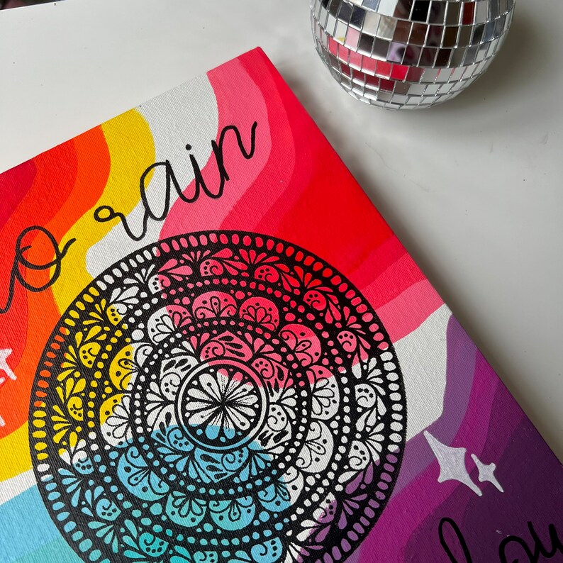 May include: A colorful canvas painting with a rainbow background and a black and white mandala in the center. The text "No rain" is written in black on the top left corner of the canvas.