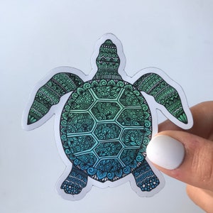 Op de afbeelding: Een gedetailleerde sticker van een zeeschildpad met een verloop van blauwe en groene tinten. De schild en het lichaam van de schildpad zijn gevuld met ingewikkelde patronen en ontwerpen, wat een unieke en artistieke uitstraling creëert. De sticker wordt door een hand vastgehouden.