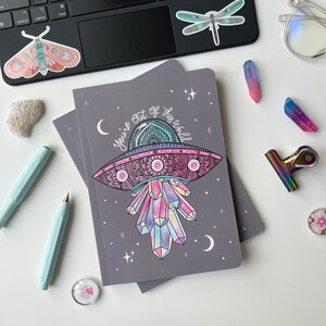 May include: A grey notebook featuring a colourful illustration of a UFO with crystals and the text "You're out of this world".
