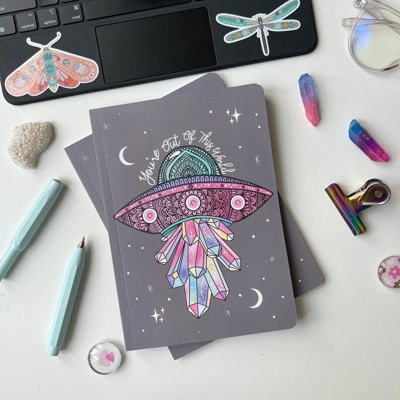 May include: A grey notebook featuring a colourful illustration of a UFO with crystals and the text "You're out of this world".
