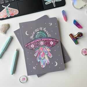 May include: A grey notebook with a colourful illustration of a UFO with crystals and the text "You're out of this world".