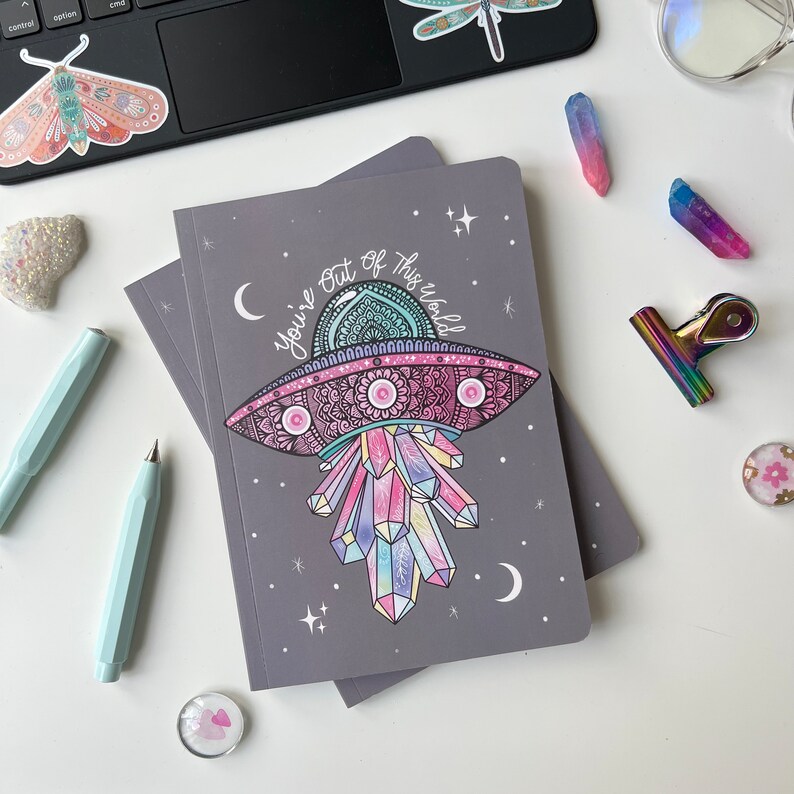 May include: A grey notebook with a colourful illustration of a UFO with crystals and the text "You're out of this world".