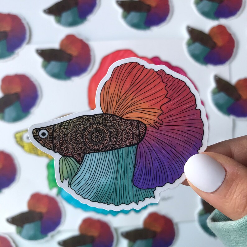 May include: A colorful sticker featuring a detailed illustration of a betta fish with a mandala pattern on its body. The fish has a red, orange, yellow, green, blue, and purple tail.