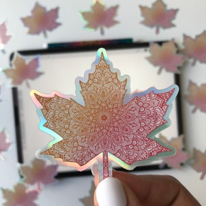 Puede incluir: Una pegatina holográfica con un diseño de mandala rojo y dorado en forma de hoja de arce.