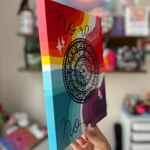 May include: A colorful canvas painting with a rainbow background and a black mandala design. The text "no rain no rainbow" is written in black cursive.