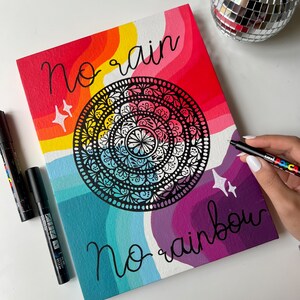 May include: A colorful canvas painting with a rainbow background and a black mandala in the center. The text "no rain no rainbow" is written in black on the canvas.