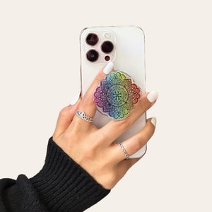 Op de afbeelding: Een witte telefoonhoes met een regenboogmandala-telefoonhouder. De telefoonhoes heeft een zwarte camera lens cover.