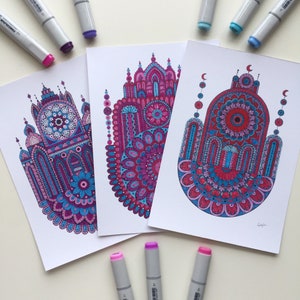 May include: Three prints of hamsa hand designs with intricate patterns in shades of pink, purple, and blue. The designs feature a central mandala and are surrounded by smaller details.