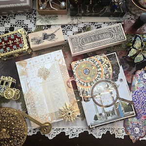 May include: An assortment of vintage-style stationery and decorative items. Includes a wooden stamp, a box labeled "LETTERS", and two illustrated cards. Gold-toned embellishments and a decorative key are also present, arranged on a lace-trimmed surface.