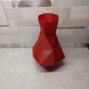 Puede incluir: Un jarrón geométrico rojo con un diseño facetado. El jarrón está hecho de un material liso y brillante.