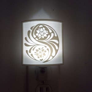 May include: White ceramic night light with a plug-in base. The light features a cut-out design of a yin yang symbol with floral accents.