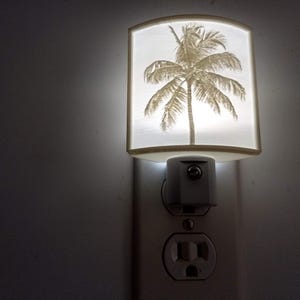 May include: A white plastic night light with a palm tree design. The night light is plugged into an electrical outlet.