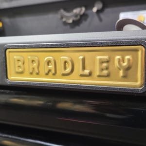 May include: A close-up of a black tool chest featuring a gold-coloured nameplate with the embossed name "BRADLEY". The nameplate is rectangular with a black border. The tool chest has black drawers and a textured surface.