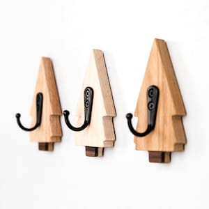 May include: Three wooden tree-shaped wall hooks with black metal hooks. The hooks are made of light and dark wood tones, with a dark wood base. The hooks are mounted on a white wall, providing a decorative and functional storage solution.
