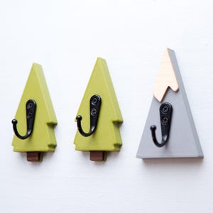 May include: Three wooden wall hooks shaped like trees and a mountain. Two are green with black hooks, and one is grey with a natural wood top and a black hook. The hooks are mounted on a white wall.