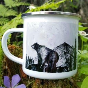 Puede incluir: Taza de camping de esmalte blanco con borde y asa plateados. La taza presenta una ilustraci&oacute;n en blanco y negro de un oso de pie frente a una cordillera y &aacute;rboles. La taza est&aacute; rodeada de follaje verde y flores.