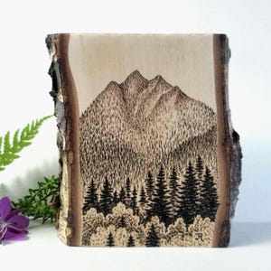 May include: A rectangular piece of light-colored wood with natural bark edges featuring a detailed black pyrography illustration of a mountain range rising above a dense forest of pine trees. The initials JR are burned into the bottom right corner.