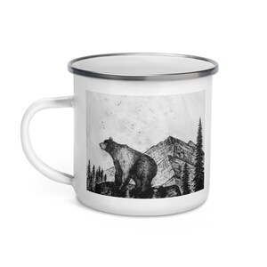 Puede incluir: Taza de esmalte blanco con una ilustraci&oacute;n en blanco y negro de un oso sobre una roca, con monta&ntilde;as y &aacute;rboles en el fondo. La taza tiene un borde plateado y un asa blanca.