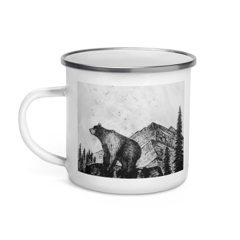 Puede incluir: Taza de esmalte blanco con una ilustraci&oacute;n en blanco y negro de un oso sobre una roca, con monta&ntilde;as y &aacute;rboles en el fondo. La taza tiene un borde plateado y un asa blanca.