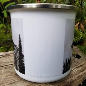 Puede incluir: Taza de esmalte blanco con borde plateado, con una escena de monta&ntilde;a y bosque en blanco y negro. La taza tiene el texto "J. Ricks Sit River Artistry Alaska" en la parte inferior.