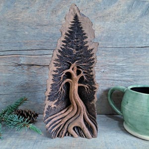 May include: A carved wooden art piece depicting a tree with a detailed root system and a full canopy of needles. The artwork is primarily brown and black, with a natural wood grain texture. The piece is displayed on a wooden surface.