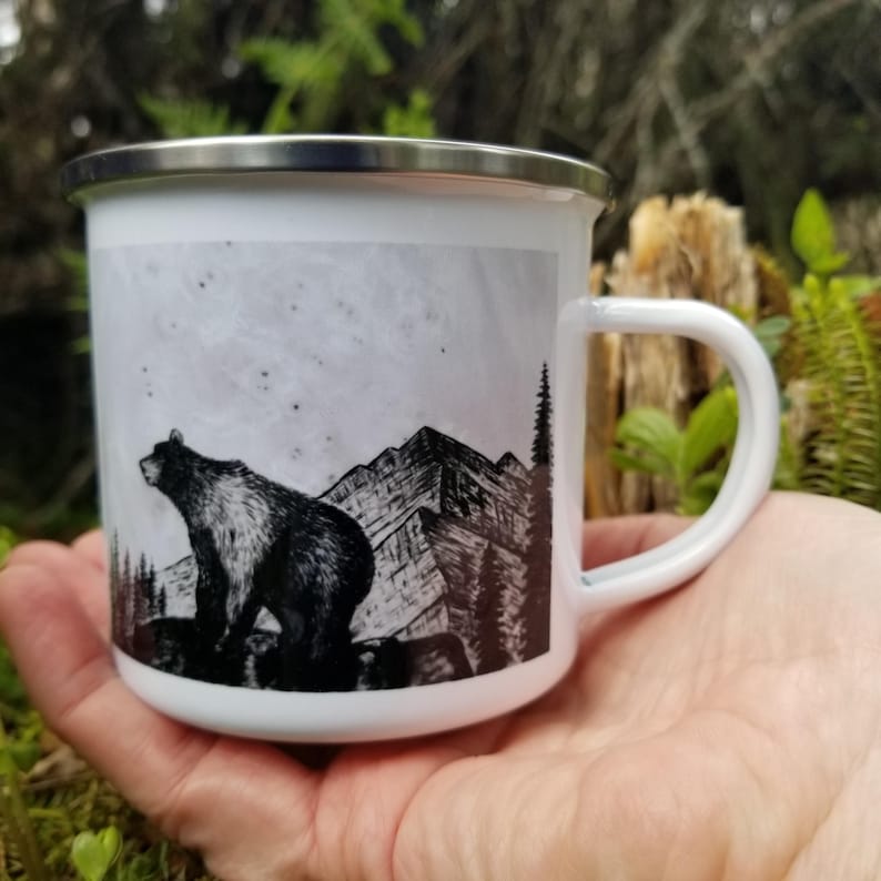 Puede incluir: Taza de esmalte blanco con una ilustraci&oacute;n en blanco y negro de un oso, monta&ntilde;as y &aacute;rboles. La taza tiene un borde plateado y un asa blanca. El dise&ntilde;o est&aacute; sobre un cielo nublado.