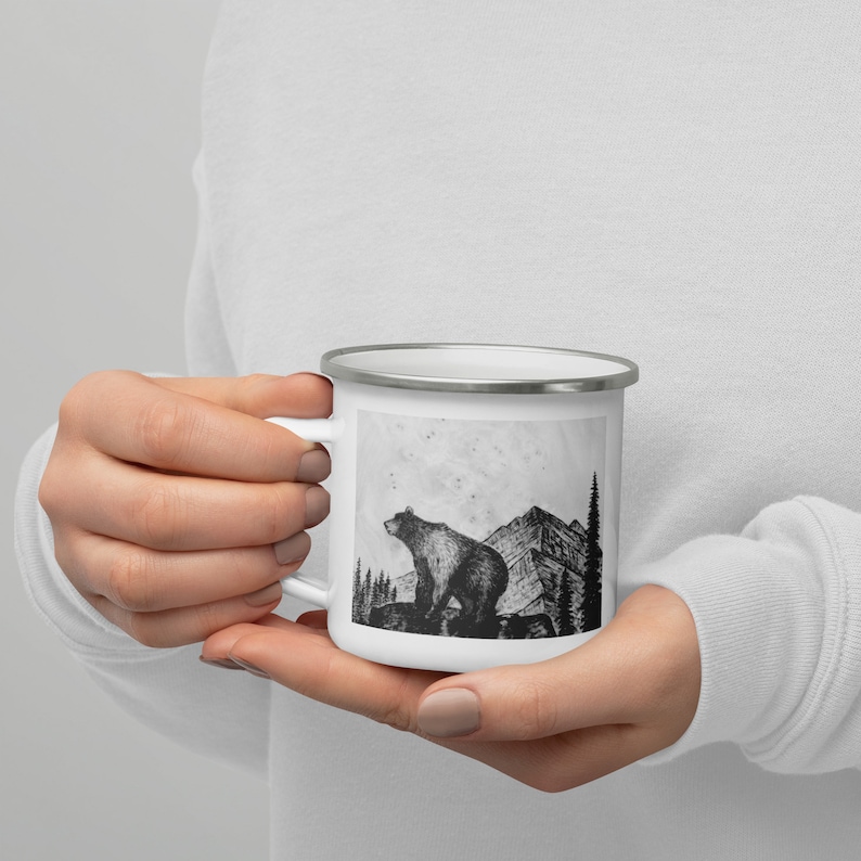 Puede incluir: Taza de esmalte blanco con una ilustraci&oacute;n en blanco y negro de un oso de pie sobre una roca, con monta&ntilde;as y &aacute;rboles en el fondo. La taza tiene un borde plateado y un asa blanca.