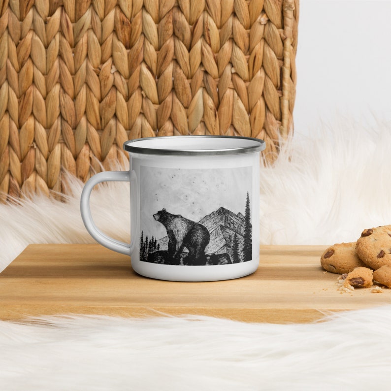 Puede incluir: Taza de esmalte blanco con una ilustraci&oacute;n en blanco y negro de un oso frente a monta&ntilde;as y &aacute;rboles. La taza tiene un borde y asa plateados. Ideal para bebidas calientes.
