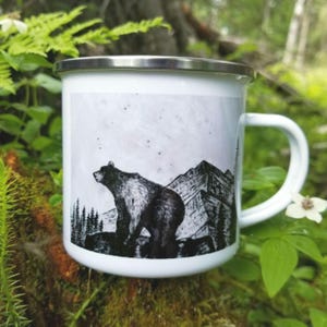A white enamel camping mug with a black and white greyscale art illustration print of a grizzly bear standing on a rock ledge in front of a mountain range. The collectable mug has a silver rim and is placed amongst forest floor ferns, and natural moss for the photo. The drawing is printed from the original hand woodburned sculpture- " Rock Ledge Bear"  by Alaska artist: J. Riske
