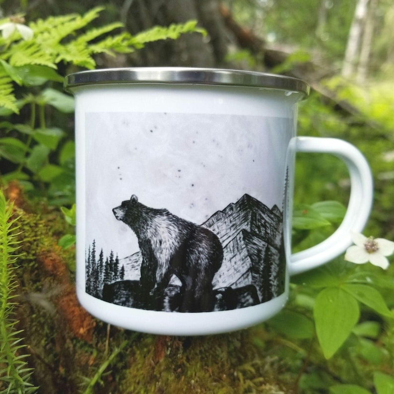 A white enamel camping mug with a black and white greyscale art illustration print of a grizzly bear standing on a rock ledge in front of a mountain range. The collectable mug has a silver rim and is placed amongst forest floor ferns, and natural moss for the photo. The drawing is printed from the original hand woodburned sculpture- " Rock Ledge Bear"  by Alaska artist: J. Riske