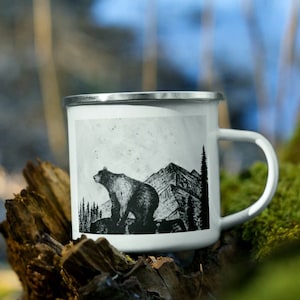A white enamel camping mug with a black and white art illustration of a grizzly bear standing on a rock ledge in front of a mountain range. The mug has a handle & a silver rim. Art by Alaska artist: J. Riske