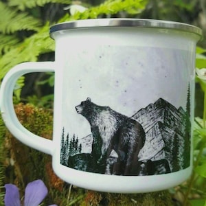 A white enamel camping mug with a black and white art illustration of a grizzly bear standing on a rock ledge in front of a mountain range. The mug has a silver rim and is placed amongst forest floor ferns, moss & blue wildflowers for the photo. Art by Alaska artist: J. Riske