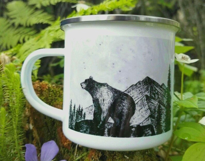 A white enamel camping mug with a black and white art illustration of a grizzly bear standing on a rock ledge in front of a mountain range. The mug has a silver rim and is placed amongst forest floor ferns, moss & blue wildflowers for the photo. Art by Alaska artist: J. Riske